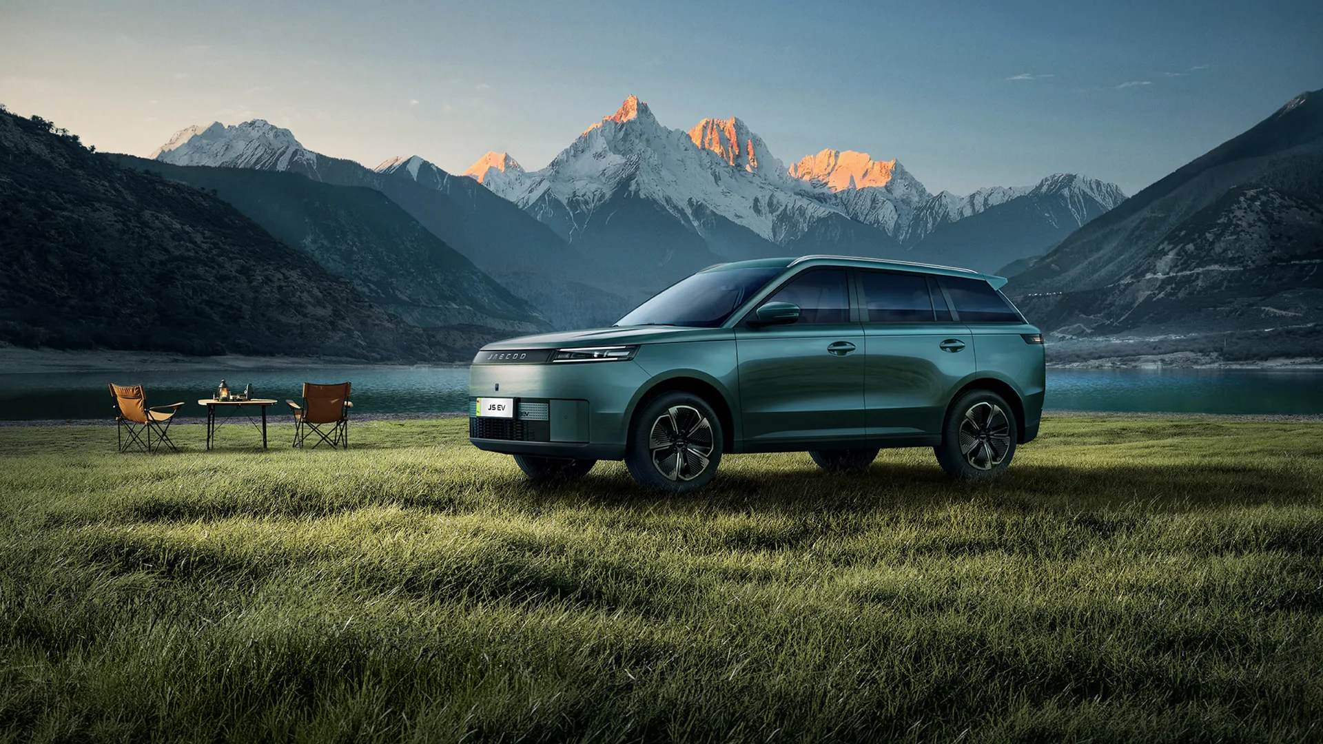 Jaecoo J5 EV in Alpine Green parked on a grass lakeshore at sunrise, snow-capped mountains behind.
